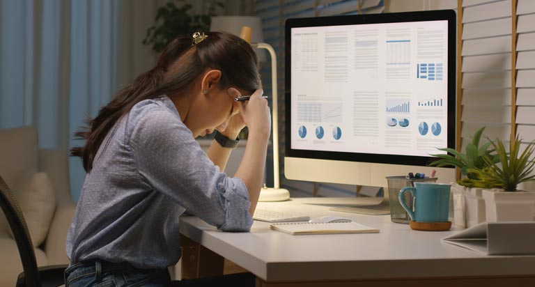 A woman sits at a desk with a computer. She’s distraught, with her elbows on the desk and her head in her hands.